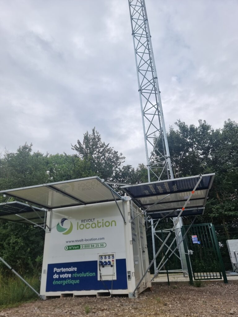 Zoom sur un container d'énergie hybride Revolt Location avec panneaux solaires photovoltaïques déployés, installé à côté d'un pylône de télécommunication.