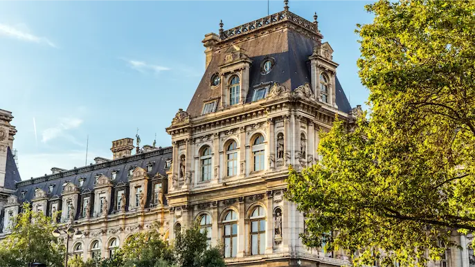 Façade historique de l'Hôtel de Ville de Paris sous un ciel bleu, partiellement encadrée par des arbres verts, illustrant un site événementiel ou une collectivité cible.