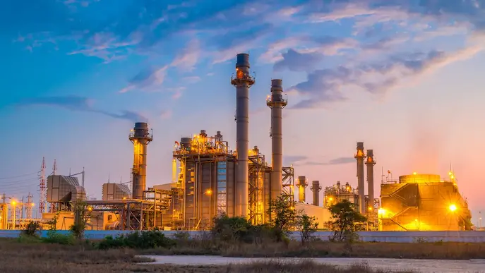 Vue panoramique d'un grand complexe industriel avec plusieurs cheminées et des installations d'éclairage jaune vif allumées au crépuscule, sous un ciel bleu et rosé.