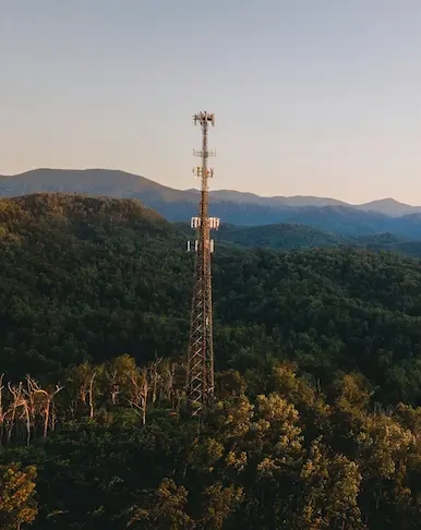 Grande antenne de télécommunication surplombant une forêt dense et vallonnée sous la lumière dorée d'un coucher de soleil.