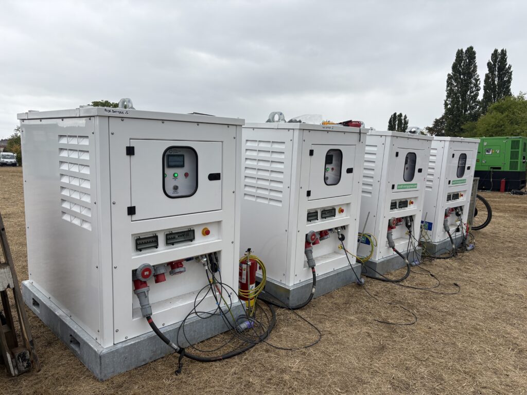 Alignement de packs batterie Revolt Energy Green dans un champ, câblés ensemble avec un groupe électrogène en arrière-plan