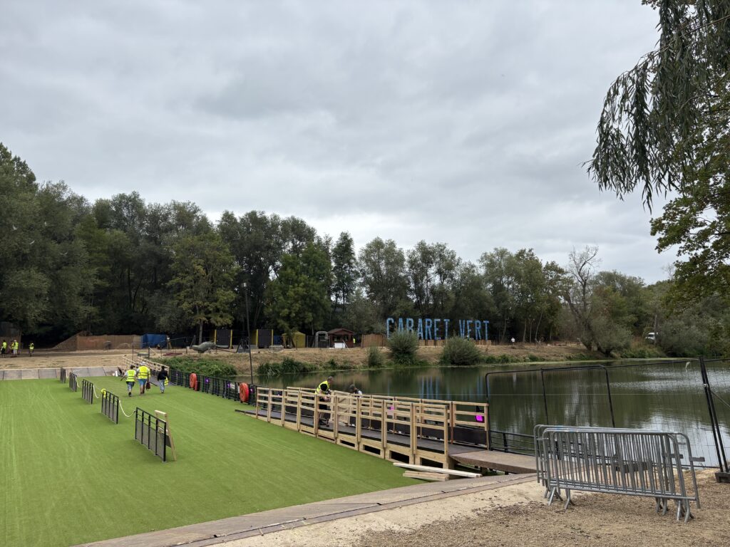 Passerelle et pelouse synthétique en cours d'installation au bord d'un plan d'eau, avec la signalétique du festival Cabaret Vert